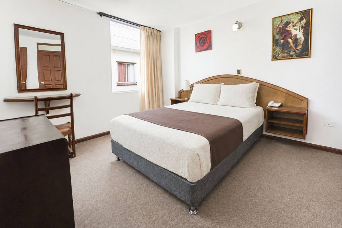 Un dormitorio con cama, mesitas de noche, escritorio, silla, ventana con cortinas y obras de arte en las paredes.