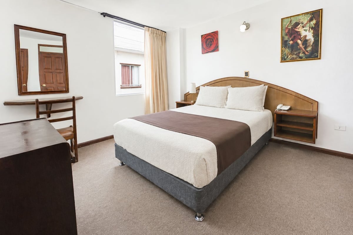 A bedroom with a bed, bedside tables, a desk, a chair, a window with curtains, and artwork on the walls.
