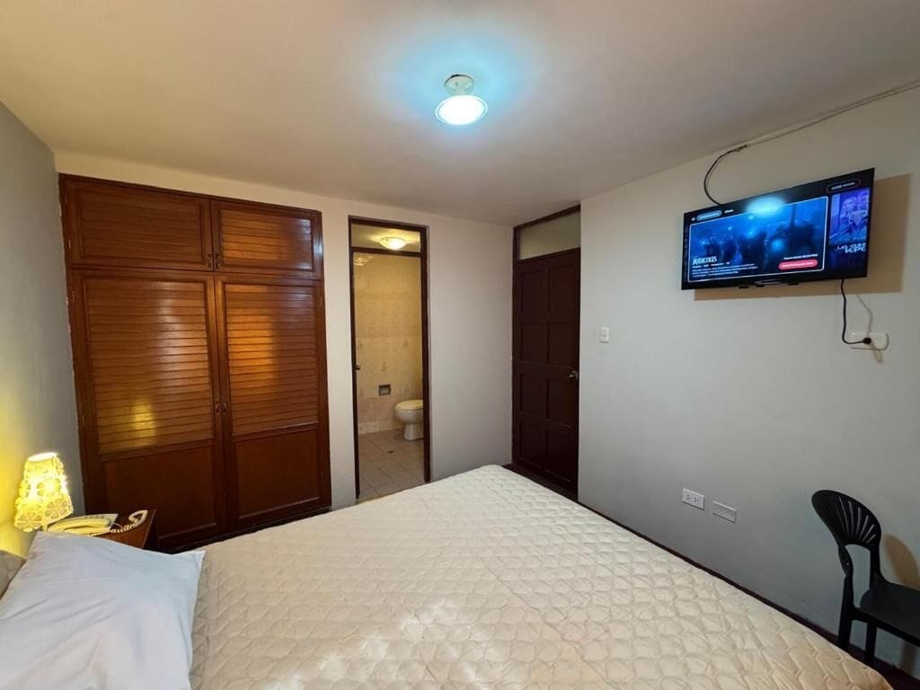 A bedroom with a bed, a television mounted on the wall, a wardrobe, and a door leading to a bathroom.