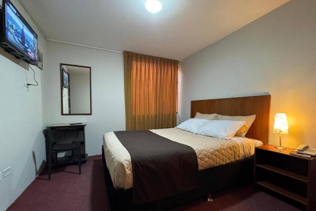 Habitación de hotel con cama, escritorio, silla, televisor, lámpara y espejo.