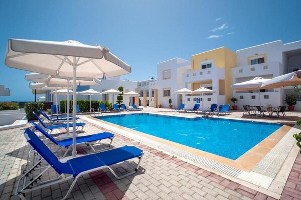 Una piscina con sillas de descanso y sombrillas, rodeada de edificios blancos con balcones.
