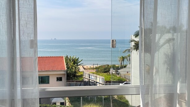 Vista a través de una ventana con cortinas translúcidas, mostrando un paisaje costero con cielo despejado, mar tranquilo y islas al distante.