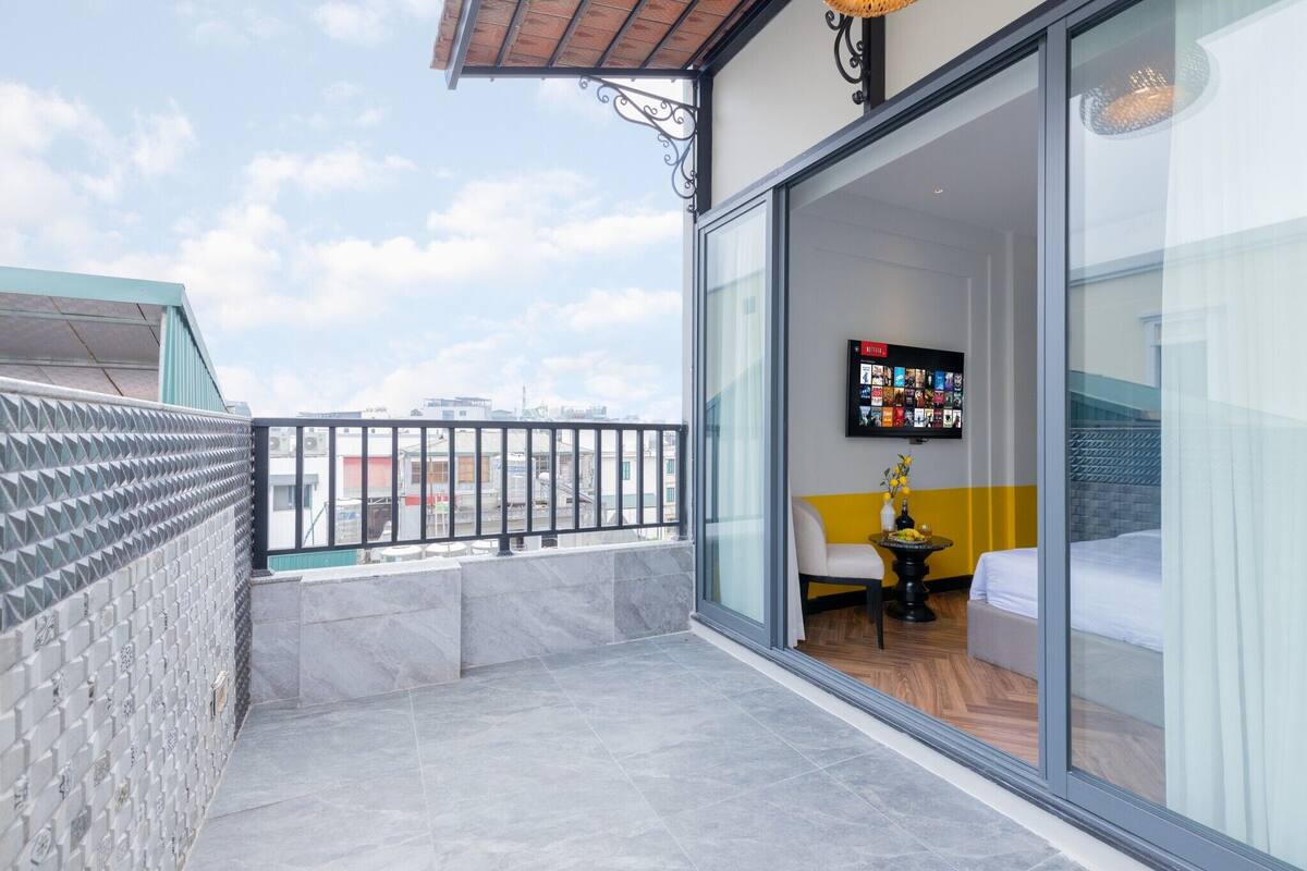 Een balkon met een betegelde vloer, een glazen deur naar een kamer met een bed, een kleine tafel met een vaas en een aan de muur bevestigde televisie.