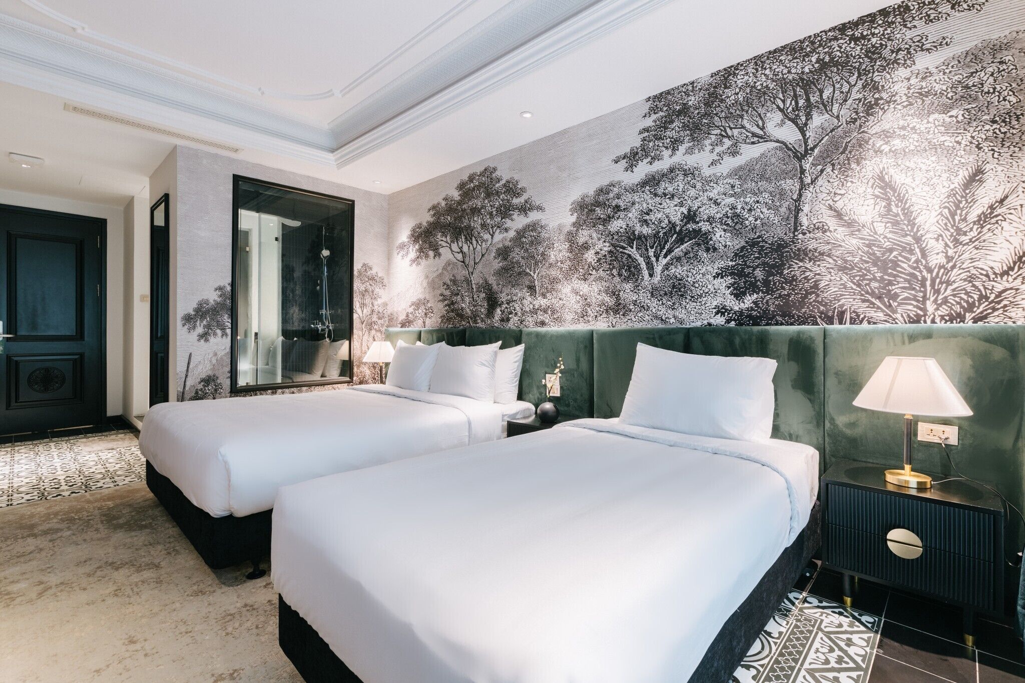 Une chambre d’hôtel avec deux lits, un grand miroir, une tête de lit verte et une fresque représentant des arbres.
