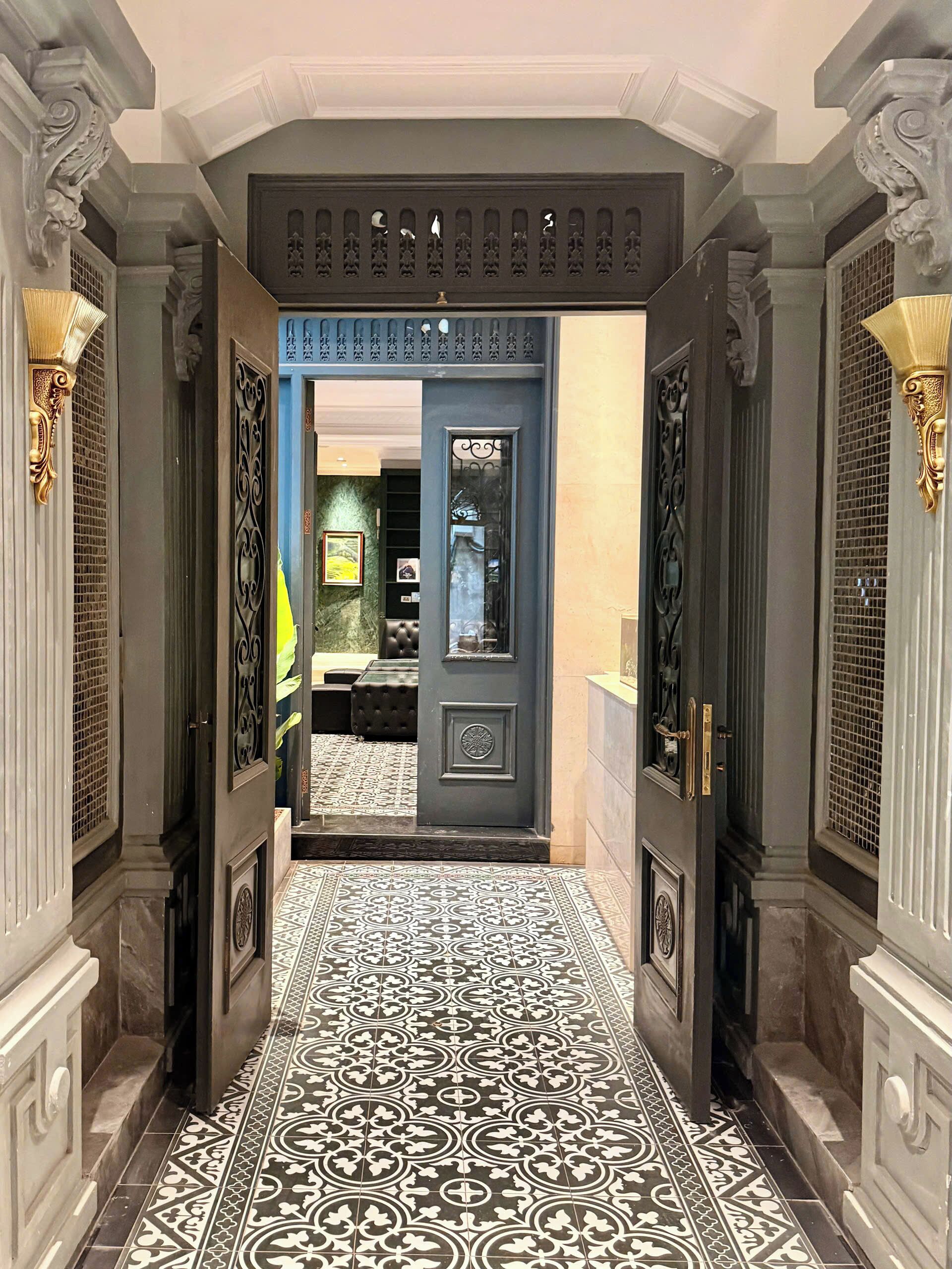A hallway with a patterned floor, a dark door with a circular emblem, and ornate wall sconces.