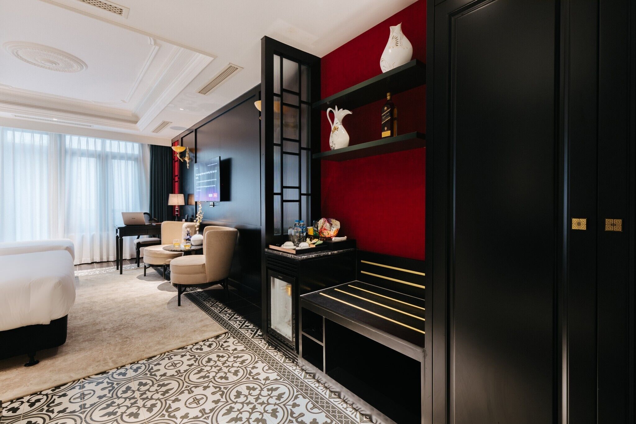 A hotel room with a bed, a desk, a chair, a sofa, a red accent wall, and a black cabinet with decorative items.