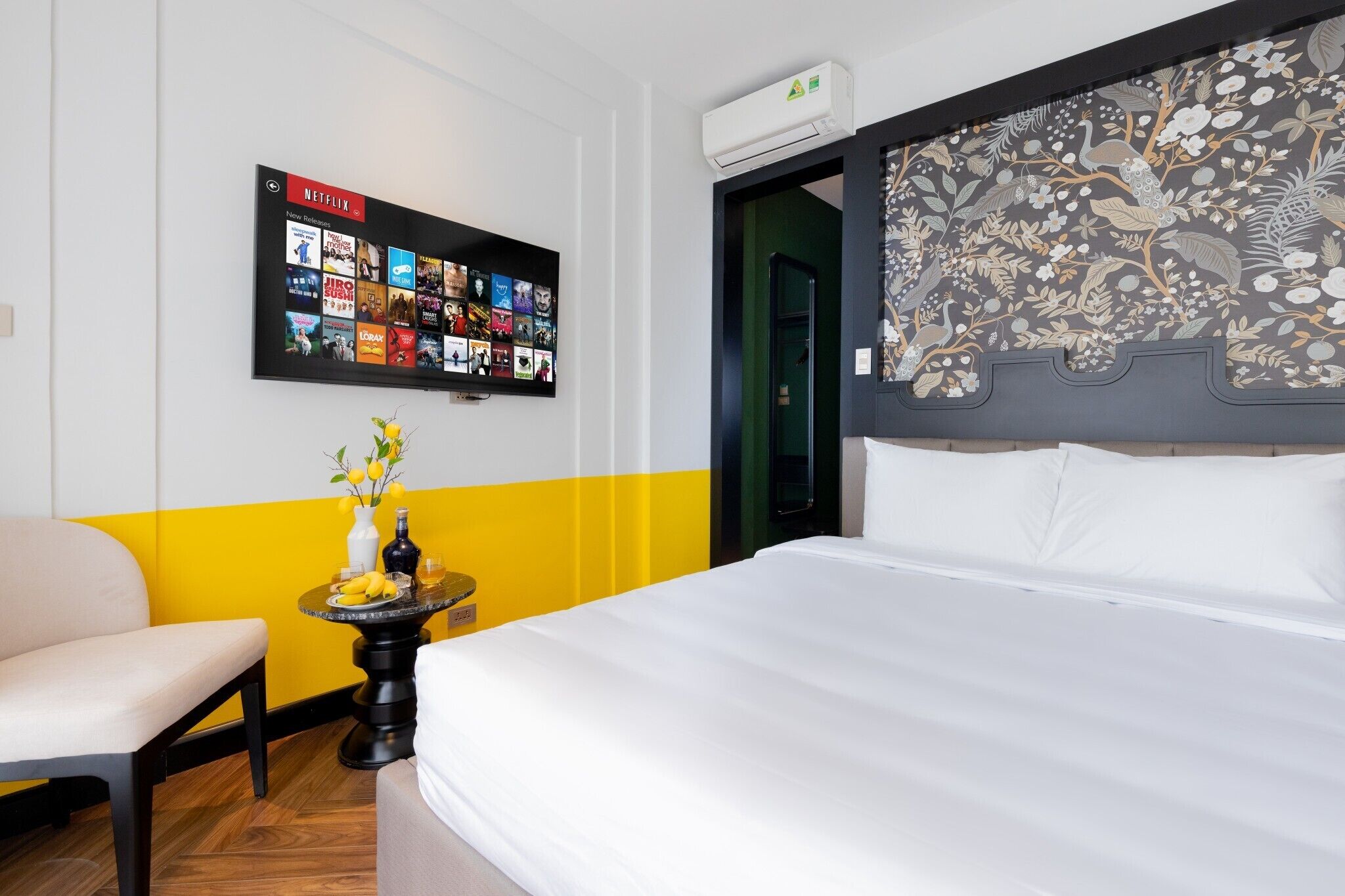 A hotel room with a bed, a wall-mounted TV displaying Netflix content, a small table with a vase and bananas, and a door with a floral design.