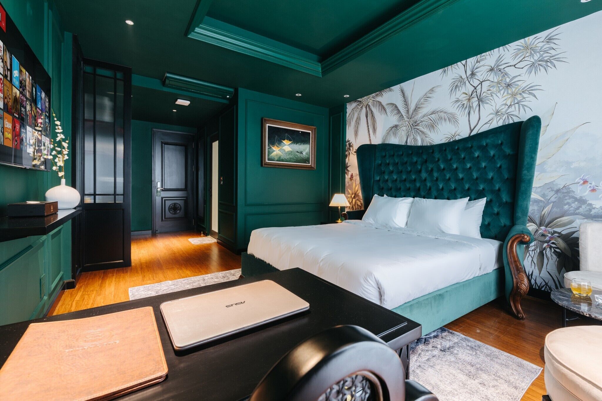 Une chambre avec un lit, un bureau, une chaise et un mur orné d’une fresque aux motifs tropicaux.