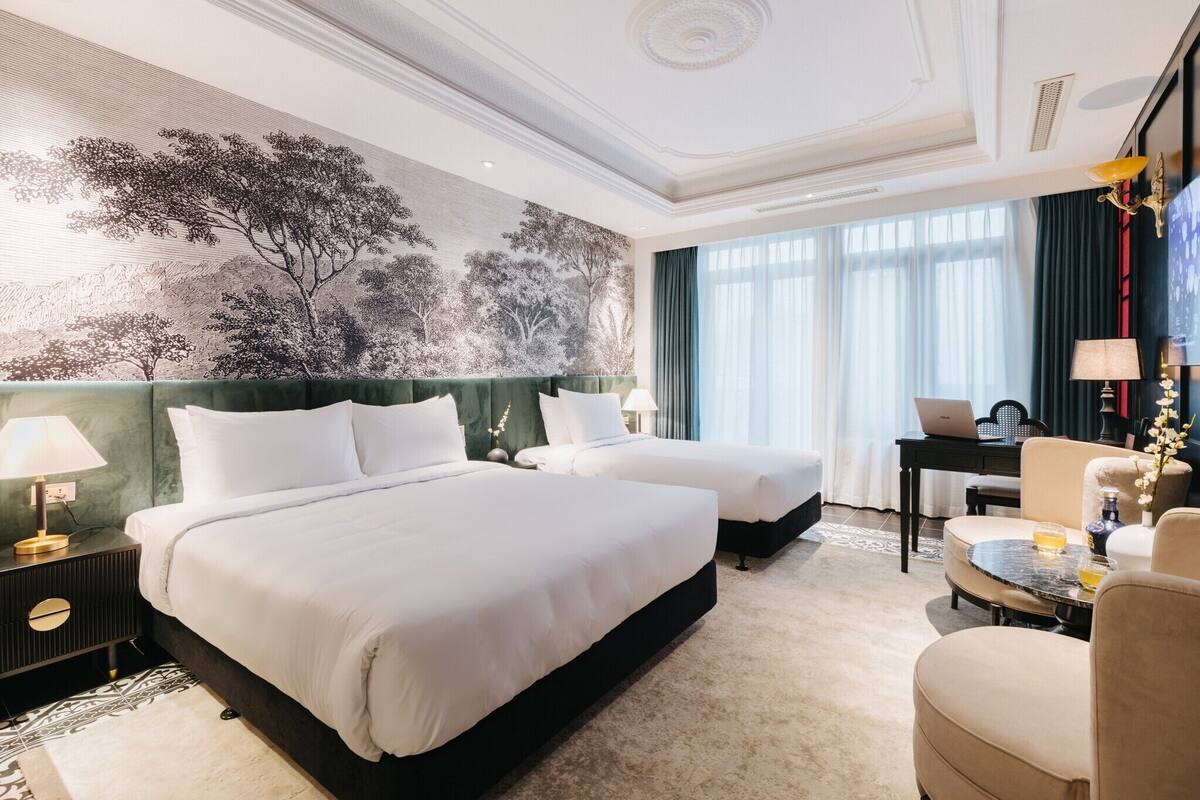 Een hotelkamer met twee bedden, een bureau met een laptop, een stoel en een groot behang met een boom.
