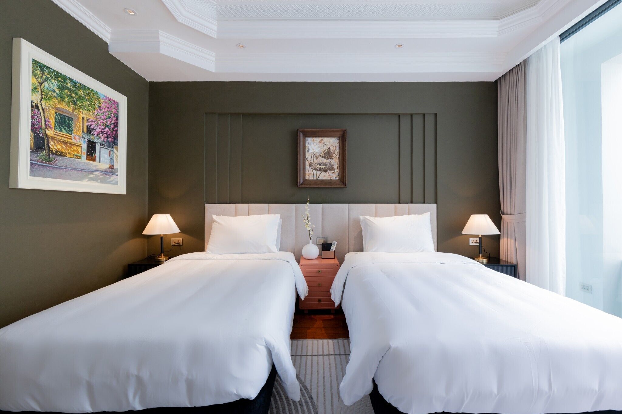 A hotel room with two beds, a nightstand, a painting, a framed picture, and a window with curtains.