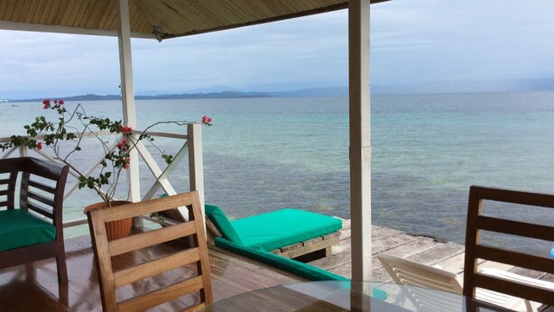 A covered outdoor seating area with wooden chairs and a table, overlooking a calm sea with distant mountains.