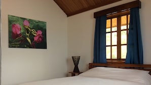 A bedroom with a bed, a window with blue curtains, a wooden headboard, and a painting of flowers on the wall.