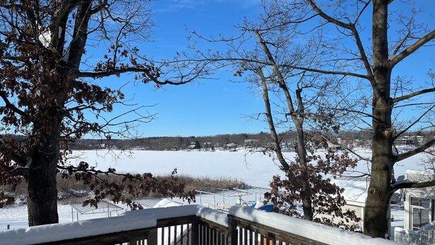 A snow-covered deck with a railing, trees, and a view of a frozen landscape.