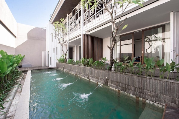 A pool area with a wooden deck, a white building with a balcony, and lush greenery. - Pelangi 148 (Seminyak)