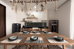 A dining area with a wooden table set for six, a kitchen in the background, and a rope chandelier. - Pelangi 148 (Seminyak)