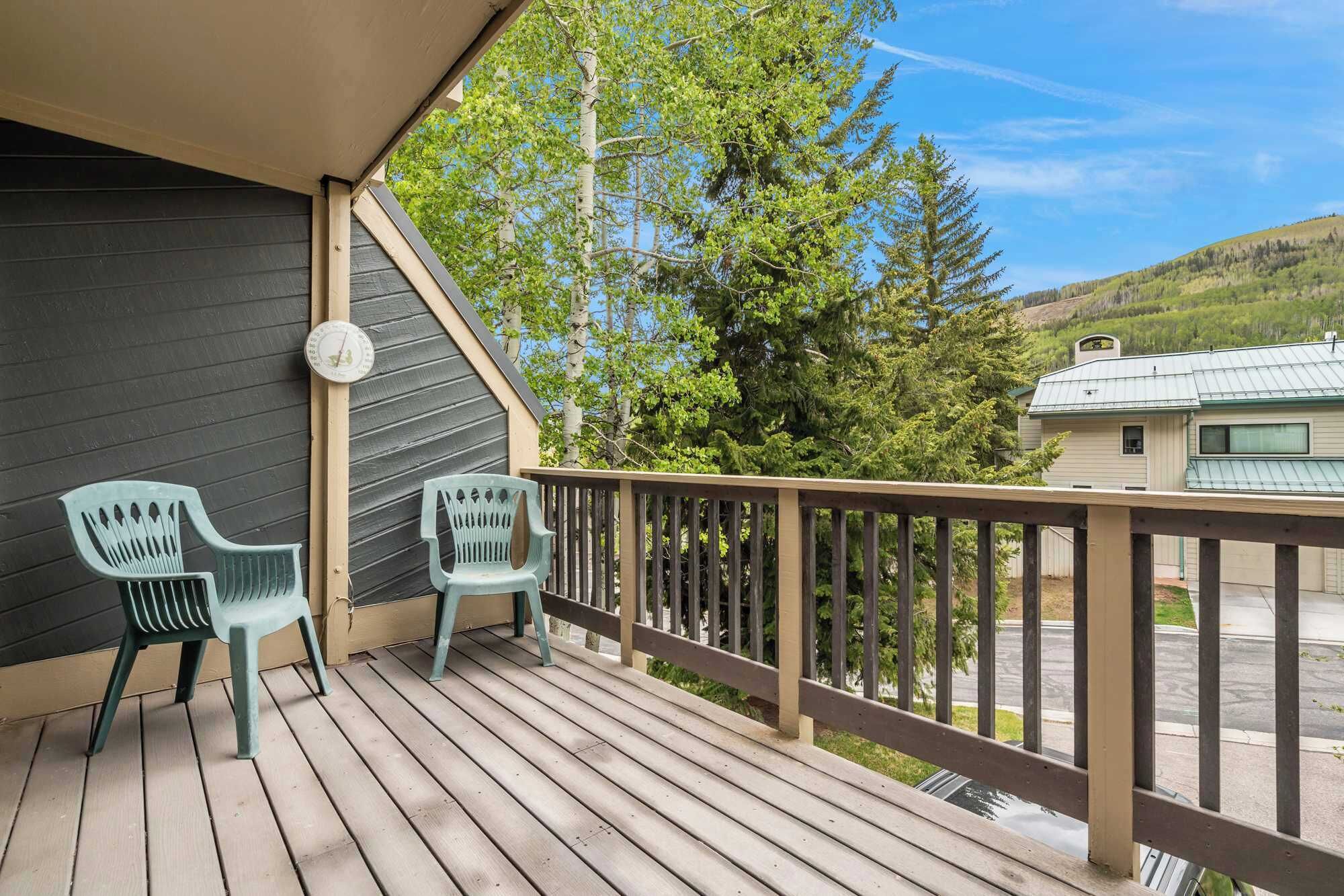 A wooden deck with two green plastic chairs, a clock, and a view of a hilly landscape with a house.