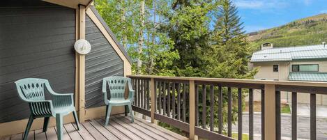 A wooden deck with two green plastic chairs, a clock, and a view of a hilly landscape with a house.