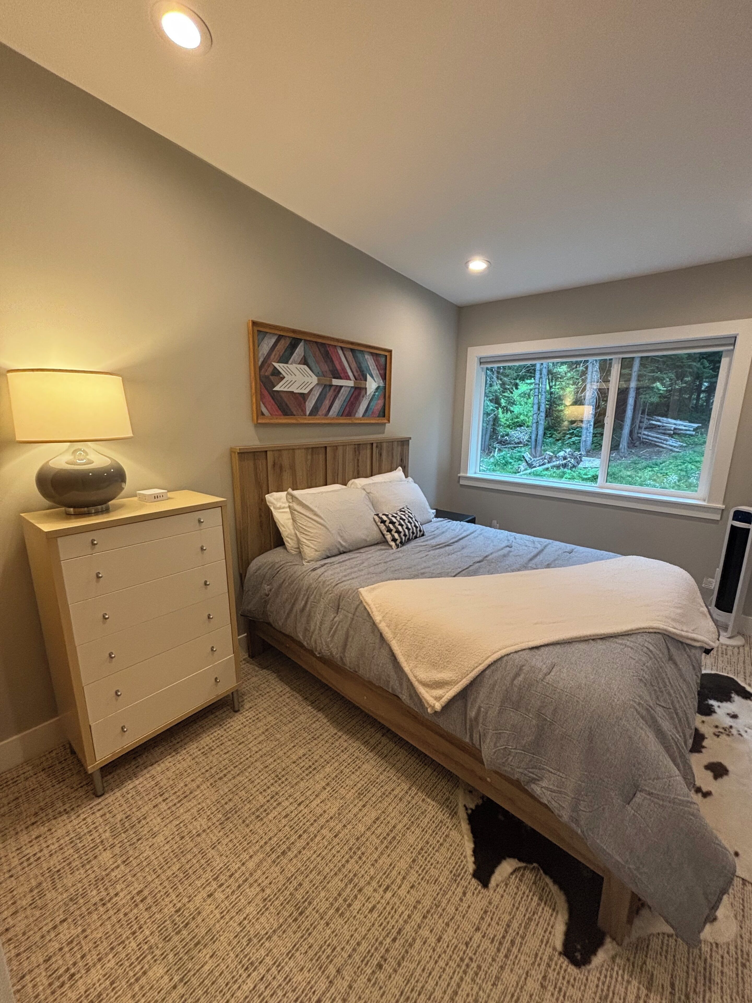 A bedroom with a bed, a dresser, a lamp, a window with a view of trees, and a wall-mounted picture.