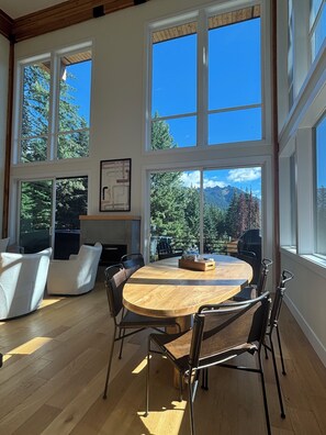 A spacious room with a large round table and chairs, large windows with a view of trees and mountains, and a wooden ceiling.