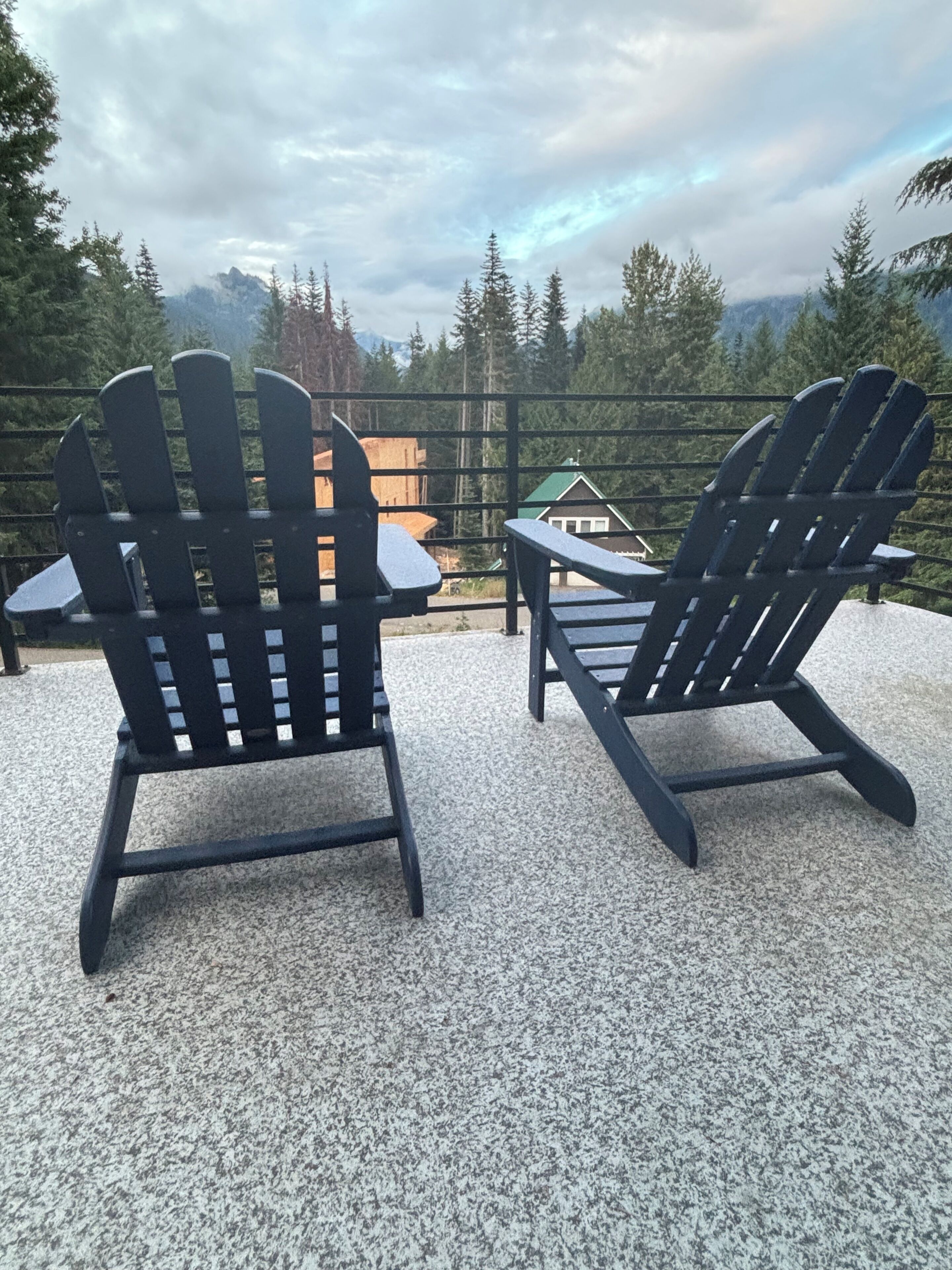 Two Adirondack chairs on a gravel surface with a view of a cabin and forest in the background.