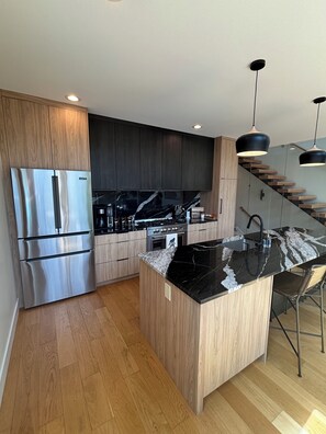 A modern kitchen with a stainless steel refrigerator, wooden cabinets, a marble countertop, and a built-in oven.