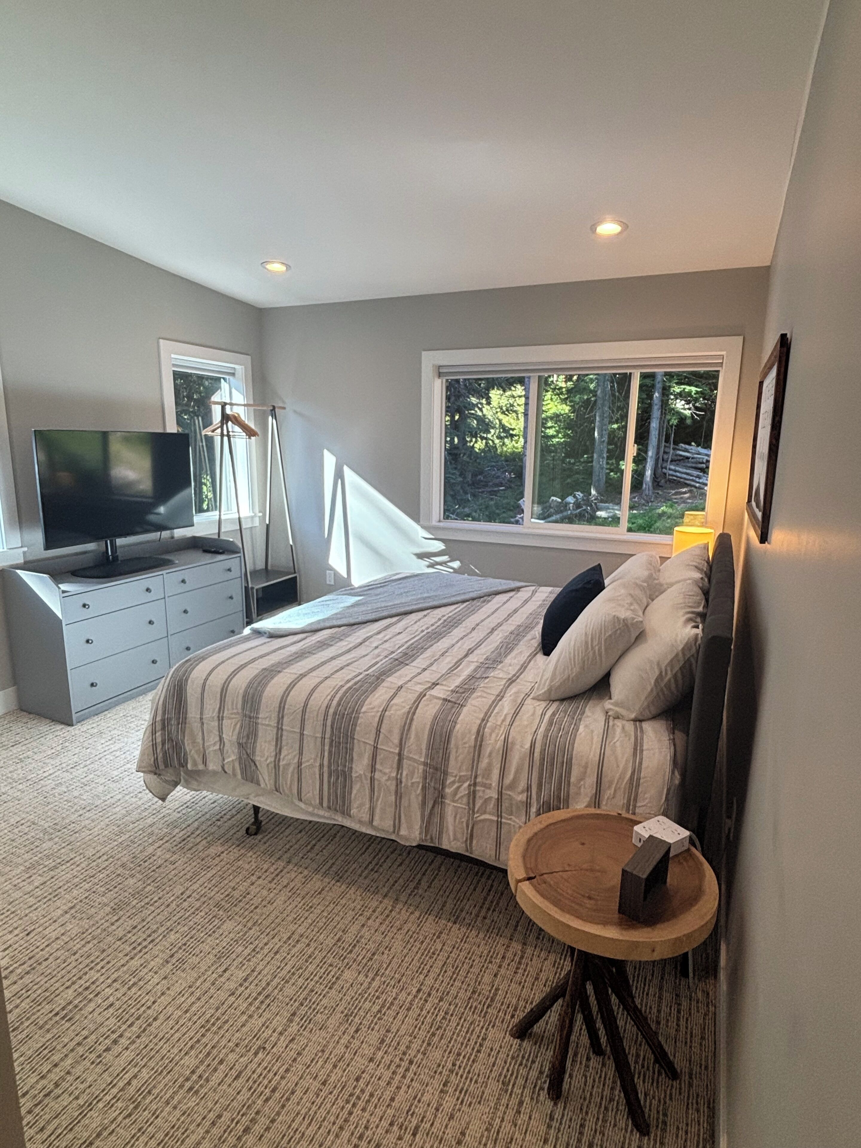 A bedroom with a bed, dresser, television, and a window with a view of trees.