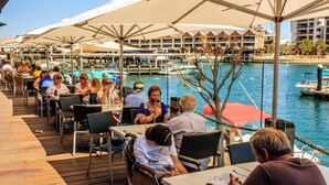 Ein Restaurant am Wasserufer mit Außenbereich, Tischen und Sonnenschirmen mit Blick auf einen Yachthafen und Boote.