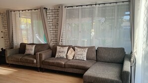 Ein modernes Wohnzimmer mit Ecksofa, einer Wand mit Backsteinoptik und transparenten Vorhängen.