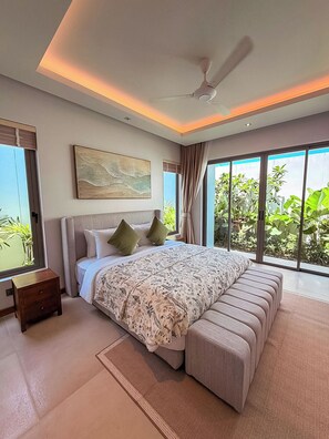 Een slaapkamer met een groot bed, een plafondventilator, een raam met uitzicht op groen en een klein nachtkastje.