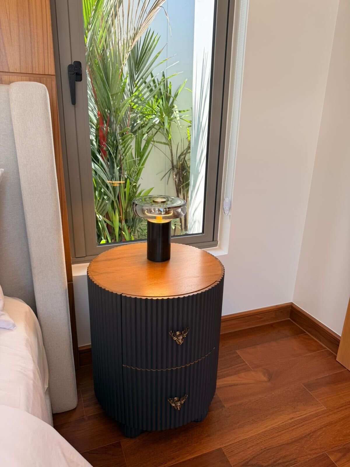 A corner room with a round, dark-colored side table with a glass top and a brass handle, a wooden floor, and a window with a view of greenery.