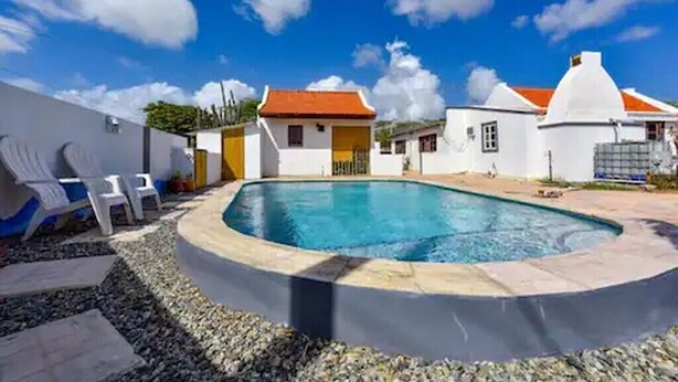 Ein Pool mit gewölbter Kante, umrahmt von einem Kiesstreifen, neben weißen Gebäuden mit orangen Dächern.
