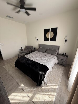Un dormitorio con cama, mesitas de noche, un tocador, un ventilador de techo y una obra de arte en la pared.