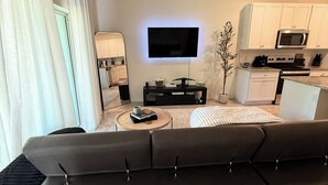 Uma sala de estar moderna com televisão de tela plana, ventilador de teto, sofá, mesa de centro e área de cozinha com armários e eletrodomésticos brancos.