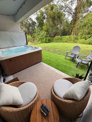 A hot tub with a wooden exterior, two wicker chairs with cushions, and a remote control on a small table.
