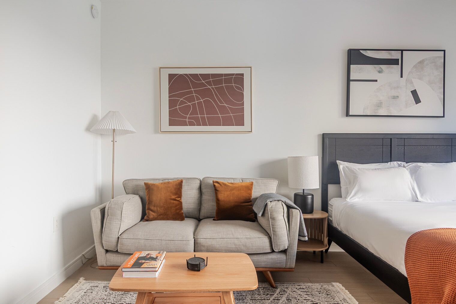 Une chambre d’hôtel moderne avec un canapé gris, une table basse en bois, une table de chevet avec une lampe et un lit avec une literie blanche.
