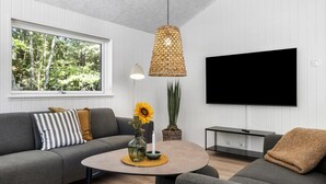 Un saló modern amb un sofà gris, una taula de centre rodona amb un vas i un girasol, una làmpada penjant de fibra natural i un televisor de pantalla plana fixat a la paret.