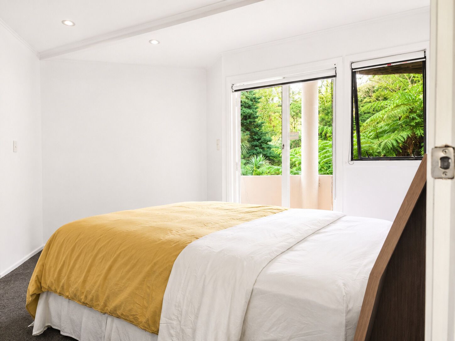 Ein Schlafzimmer mit Bett, weißen Wänden, einem Fenster mit Blick auf Grünflächen und einer Tür.