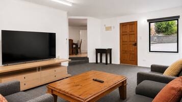 Sala de estar con televisor de pantalla plana, una mesa de centro de madera, un sofá gris y una puerta que da a un balcón con vistas a zonas verdes.