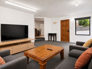 Ein Wohnzimmer mit Flachbildfernseher, einem Holztisch, einer grauen Couch und einer Tür zu einem Balkon mit Grünblick.
