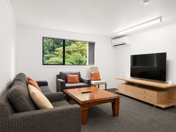 Ein modernes Wohnzimmer mit einer Couch, Sesseln, einem Couchtisch, einem Flachbildfernseher und einem großen Fenster mit Blick auf Grünflächen.