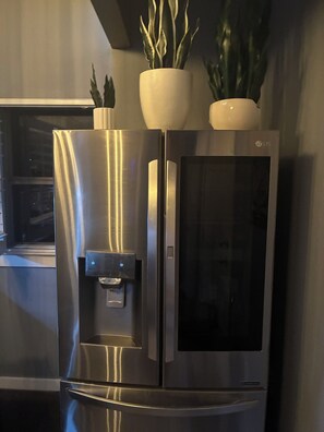 Un refrigerador de acero inoxidable con dispensador de agua, dos plantas en macetas encima y una ventana al fondo.