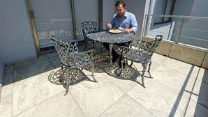 En man sitter och äter vid ett runt bord med utsmyckade metallstolar på en balkong med en glasdörr och ett metallräcke.