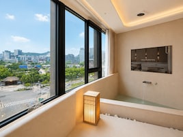 A modern bathroom with a bathtub, a window with a city view, and a wall-mounted mirror.