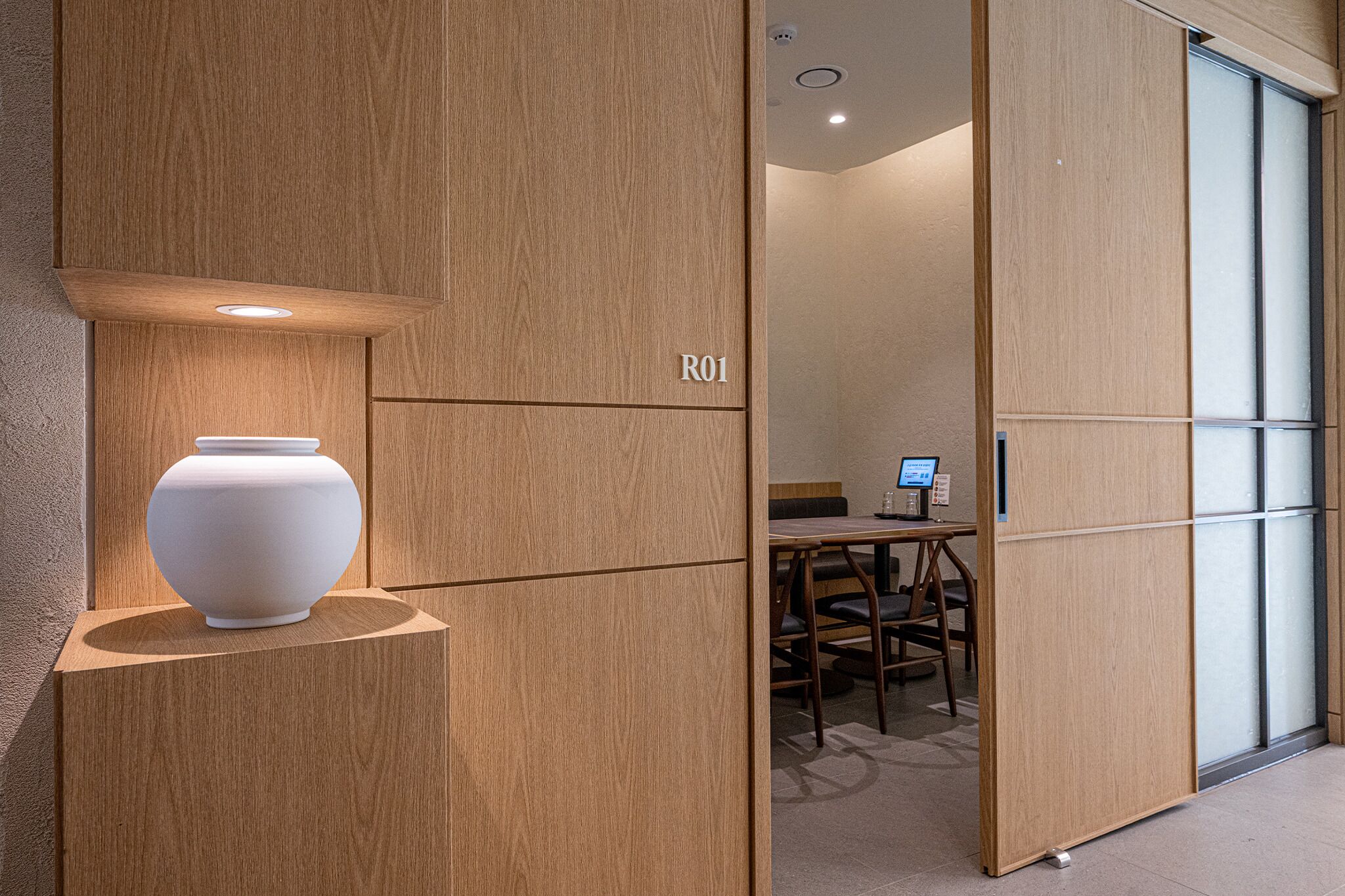 A modern interior with a wooden partition labeled 'R01', a white vase on a shelf, and a desk with a computer monitor.