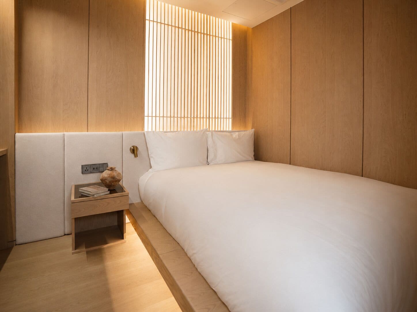 Un dormitorio con cama, mesita de noche y ventana con persianas verticales.