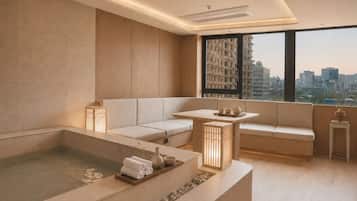 A modern spa room with a large bathtub, a sofa, a small table with tea set, and a view of the city through large windows.