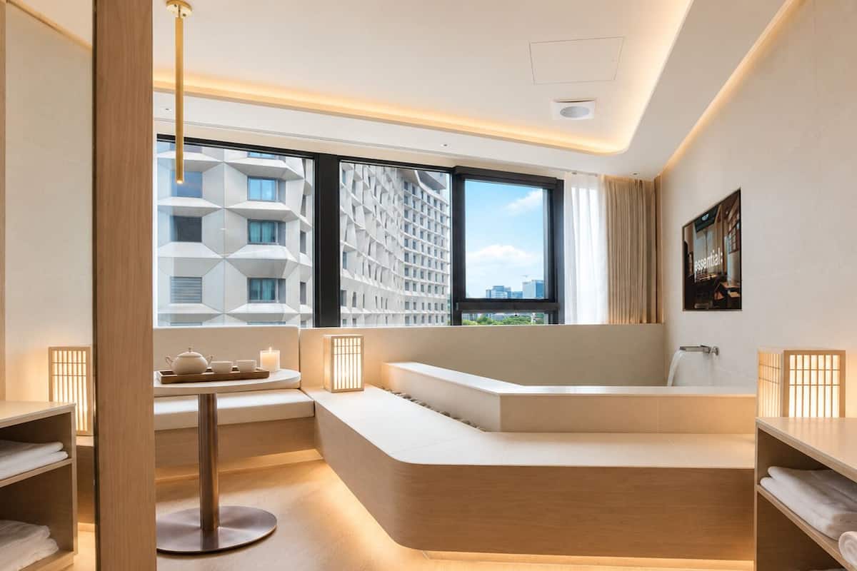 A modern hotel room with a curved seating area, a large window with a city view, a small table with a tea set, and a wall-mounted picture.
