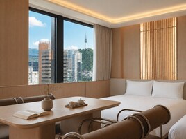 A hotel room with a large window, a bed, a seating area with a table, and a view of a cityscape.