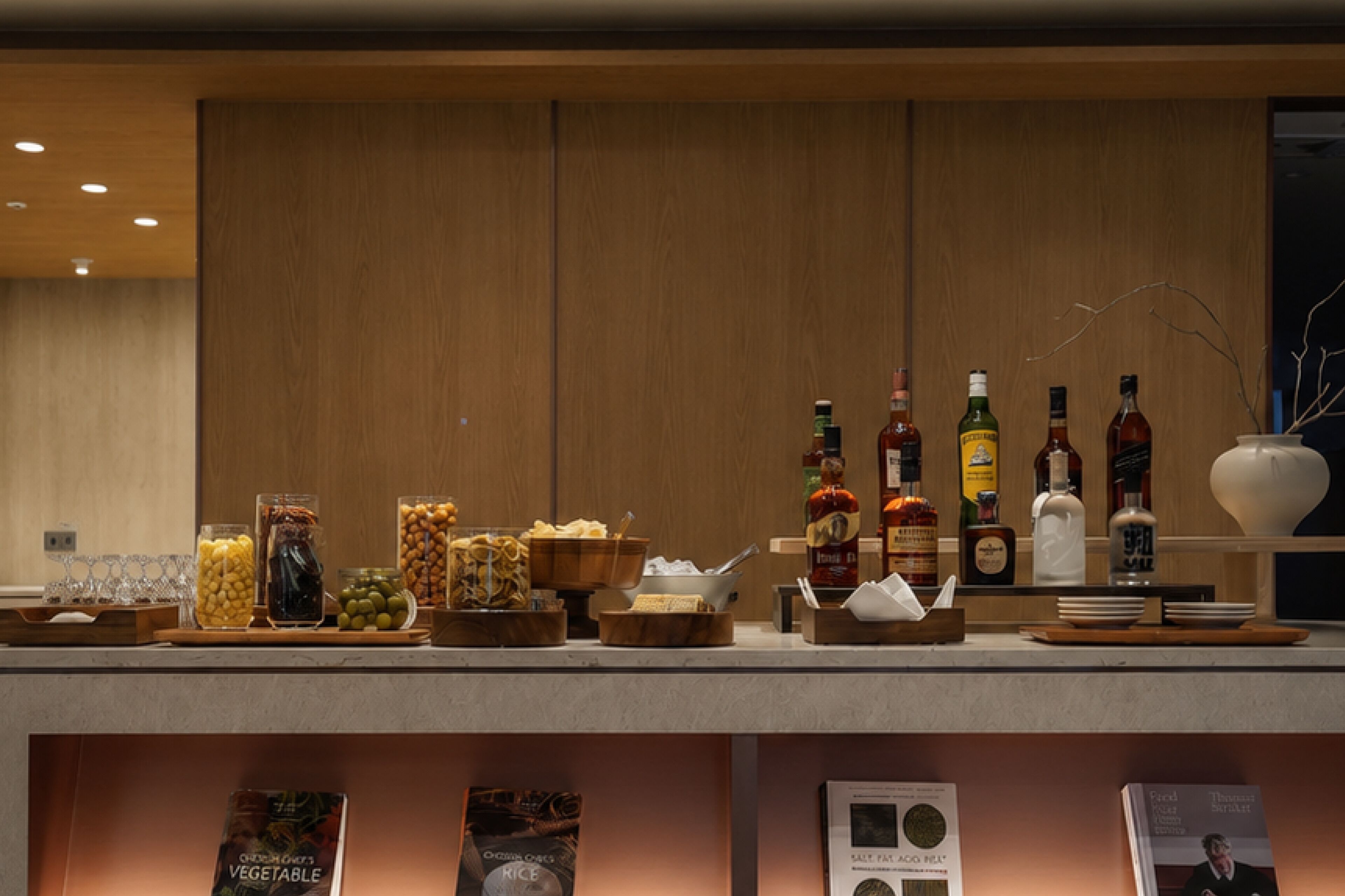 Una zona de bar con varias botellas y cristalería, un bol de aperitivos y una selección de libros en la encimera.