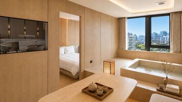 A modern hotel room with a bed, a sitting area with a tray of tea, a bathtub, and a city view through large windows.